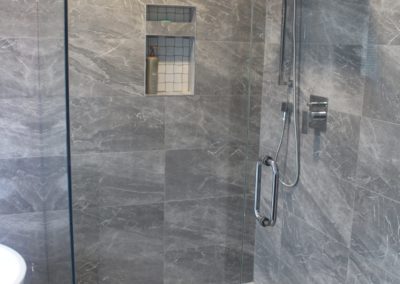 Luxurious renovated grey shower with a spa-like atmosphere. The grey tiles and glass enclosure create a sleek and modern look. Features include a rainfall showerhead and multiple body jets for the ultimate shower experience