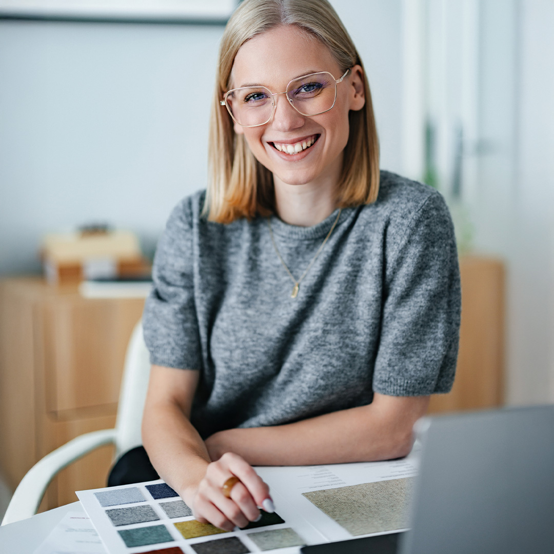 A young, professional businesswoman seated at her workspace in a design studio, reviewing material samples with a laptop.